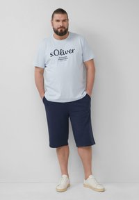 Light blue short-sleeve t-shirt with dark blue logo text, paired with navy shorts, and white sneakers. Smooth fabric, casual fit.