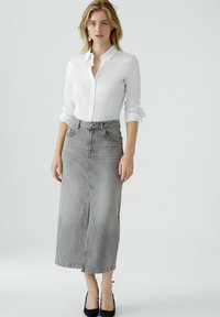 Graue Denim-Maxikleid mit einem Schlitz vorne und einem Fünf-Taschen-Design, kombiniert mit einem weißen Hemd und schwarzen hohen Absätzen.