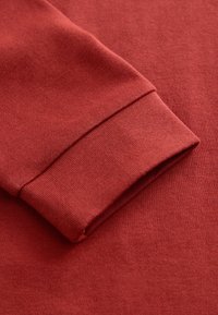 Detail složeného manžetu dlouhého rukávu na měkkém červeném oděvu s viditelnou pletenou texturou.