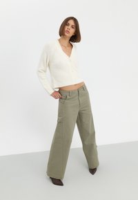 Crèmekleurige, korte cardigan met een v-hals en een zachte textuur, gecombineerd met hooglopende, wijdvallende olijfgroene broeken.