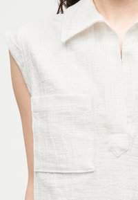 Λευκό μπλουζάκι χωρίς μανίκια με υφή, ευρύ γιακά και τσέπη στο στήθος, φορεμένο από άτομο με σκούρα μαλλιά που είναι δεμένα πίσω.