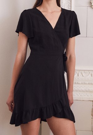 Frau trägt ein kurzes schwarzes Wickelkleid mit Rüschensaum und kurzen, flatternden Ärmeln, steht vor einer weißen, dekorativen Wand mit Fliesen.