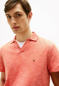 Polo shirt in cotone di un vivace color corallo con una trama testurizzata. Presenta un collo a V e un piccolo logo ricamato sul lato sinistro del petto. Maniche corte.