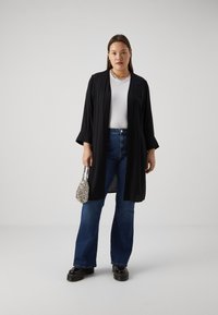 Un long kimono noir sur un t-shirt blanc, associé à un jean bleu évasé et des chaussures à plateforme noires ; tenant un petit sac à main en perles argentées.