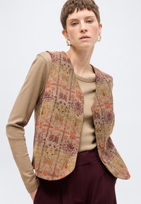 Γυναίκα με κοντά μαλλιά που φοράει σκουλαρίκια χρυσούς κρίκους, μπεζ μακρυμάνικο πουκάμισο, εμπριμέ γιλέκο με κουμπιά και σκούρο μπορντό παντελόνι, ποζάροντας σε λευκό φόντο.