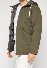Hombre vistiendo una chaqueta con capucha de color verde oliva abierta, con cordones blancos, una camiseta blanca y pantalones blancos sobre un fondo liso.