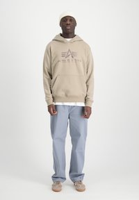 Sudadera con capucha beige con un logo, bolsillo frontal, sobre una camiseta blanca. Unos pantalones azul claros y zapatillas color arena completan el conjunto.