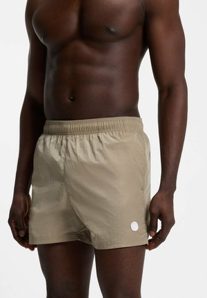 Muskuløs mørkhudet mand iført beige badeshorts med elastisk talje og lille hvidt cirkulært logo på venstre ben.