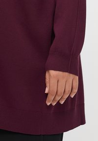 Burgundy knitted long-sleeve top with ribbed cuffs, displaying a soft texture and straight hemline, worn over black pants.