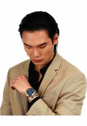 Sølv- og blå armbåndsur med en rund urskive og metalband, båret på en hånd. Beige blazer over en sort skjorte.