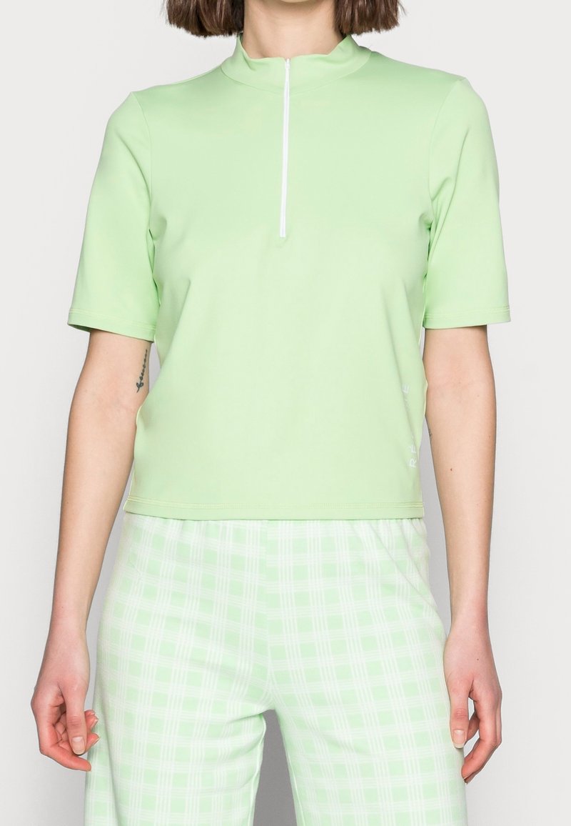 Haut à manches courtes et col montant en tissu vert clair, avec une fermeture éclair à l'avant et une coupe décontractée, associé à un pantalon à carreaux verts.