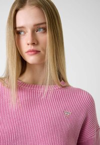Knitted pink sweater with a ribbed texture, featuring a small triangular logo patch on the left chest. Round neckline, relaxed fit.