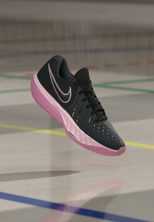 Czarno-różowy sportowy but Nike unoszący się nad halowym boiskiem z widocznymi na podłodze kolorowymi liniami granicznymi.