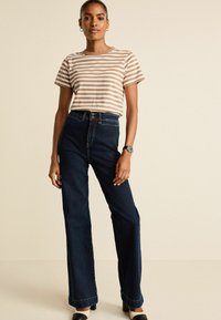 Gestreiftes braunes und weißes T-Shirt, hochtaillierte dunkelblaue Jeans, slim fit, glatte Textur, kombiniert mit schwarzen und weißen Blockabsatzschuhen.