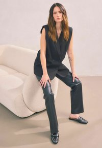 Svart dubbelknäppt ärmlös blazer, kombinerad med åtsittande svarta läderbyxor och nitarbärande svarta loafers. Mjuk, beige texturerad soffa i bakgrunden.