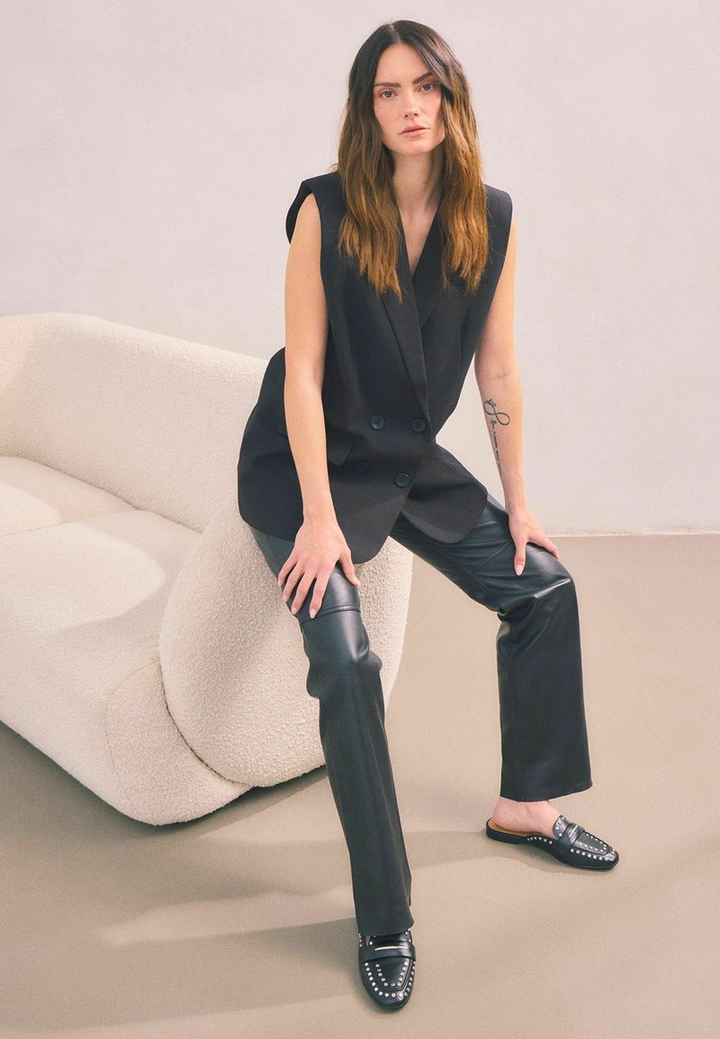 Svart dubbelknäppt ärmlös blazer, kombinerad med åtsittande svarta läderbyxor och nitarbärande svarta loafers. Mjuk, beige texturerad soffa i bakgrunden.