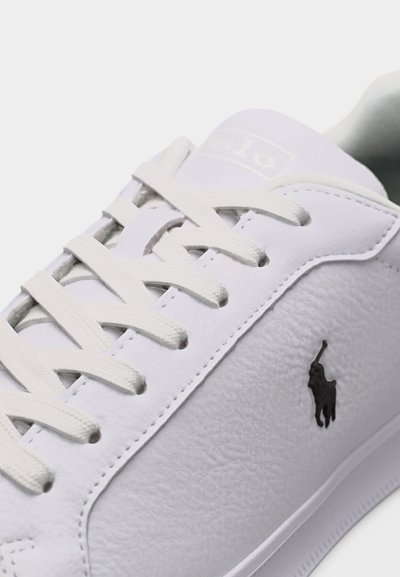 Λευκό δερμάτινο αθλητικό παπούτσι με λευκά κορδόνια και μαύρο λογότυπο παίκτη πόλο στο πλάι, εμφανιζόμενο σε κοντινή γωνία.