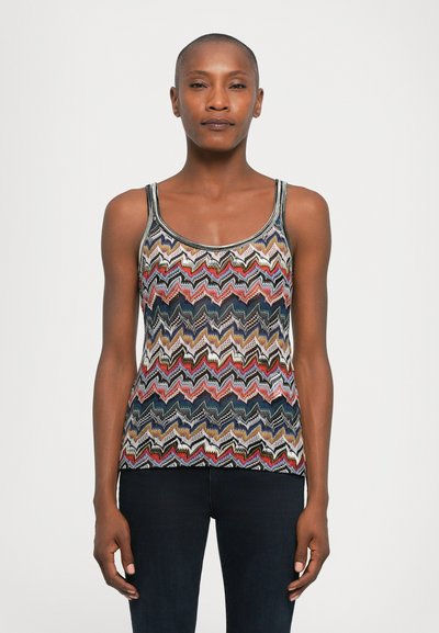 Vrouw in een mouwloze top met een meerkleurig zigzagpatroon en donkere broek, staand tegen een effen lichte achtergrond.