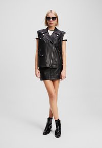 Gilet en cuir noir avec épaulettes, fermeture éclair et poches, associé à une jupe en cuir noir ajustée et des bottines noires à talons hauts.
