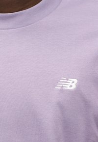 Ανοιχτό μωβ βαμβακερό μπλουζάκι με στρογγυλή λαιμόκοψη και λευκό κεντημένο λογότυπο στην κάτω δεξιά πλευρά. Μαλακή υφή και απλός σχεδιασμός.