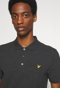 Donkergrijze polo-shirt met een textuurstof, met een gele geborduurde logo op de borst en drie knopen bij de kraag.