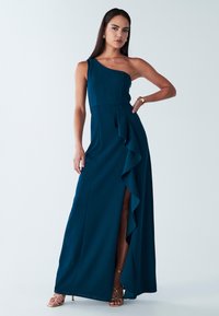 Mujer con un vestido de noche asimétrico color verde azulado con una abertura con volantes hasta el muslo, pendientes de aro dorados, reloj y sandalias doradas de tiras.