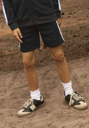 Svart zip-up jacka och matchande shorts med vita sidostråkar. Bär svart och beige sneakers med vita strumpor. Mark är jord.