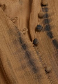 Tissu marron avec une finition texturée, agrémenté de boutons ronds proéminents et d'accents noirs subtils. Le matériau semble léger et légèrement froissé.