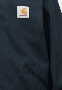 Granatowa kurtka Carhartt z przeszywaną naszywką z pomarańczową literą „C” i skośną kieszenią z przodu.