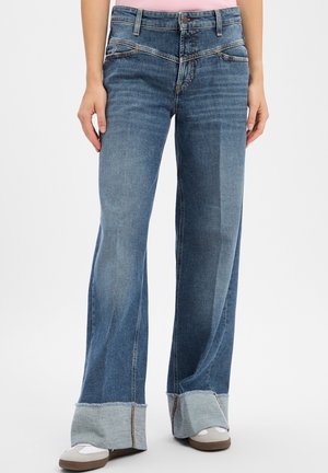 Mittblau gewaschene Jeans mit weitem Bein und großen umgeschlagenen Säumen, getragen mit lässigen weißen und beigen Sneakern.