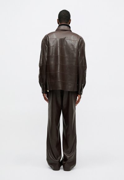 Egy férfi áll, háttal fordulva, sötétbarna bőrkabátot és hozzá illő bővebb bőr nadrágot viselve egy egyszínű háttér előtt.