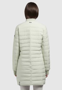 Cămașă căptușită de culoare verde deschis, cu o formă lungă și rectangulară. Prezintă cusături orizontale quilted, un guler înalt și mansete elastice la mâneci.