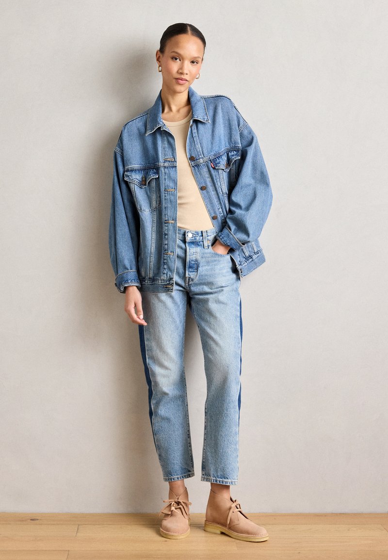 Giacca di jeans azzurro chiaro con vestibilità comoda, top beige, jeans azzurro chiaro con strisce scure sui lati e stivaletti beige.