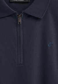 Marinblå polo skorta med dragkedja och krage, med en subtil broderad logotyp på bröstet. Tillverkad av mjukt, texturerat bomullstyg.