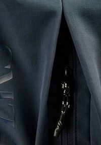 Zbliżenie ciemnoniebieskiej tkaniny z subtelnym wzorem kratki, częściowo zakrywającej czarny zamek błyskawiczny z plecionym sznurkiem do ściągania.