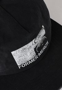 Black cap with textured fabric featuring a white graphic and text "FORMER MERCH." Design includes a rectangular shape with curved brim.
