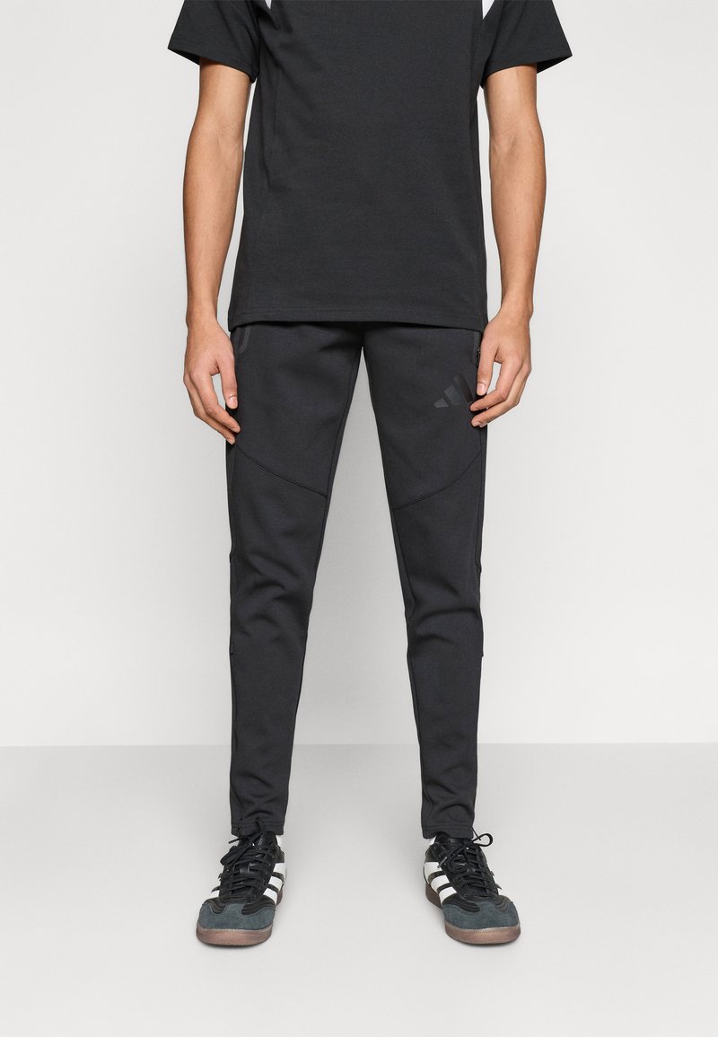 Μαύρο αθλητικό σύνολο που περιλαμβάνει σκούρο μπλουζάκι και μαύρο παντελόνι στενής εφαρμογής με τσέπες με φερμουάρ και υφασμάτινες λεπτομέρειες. Μαύρα και λευκά παπούτσια.