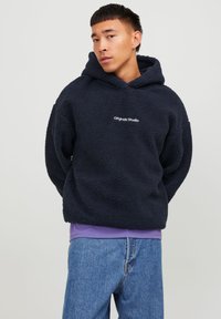 Navy blue fleece hoodie with a textured surface and a kangaroo pocket, embroidered "Originals Studio" logo on the chest. Worn over a purple shirt.