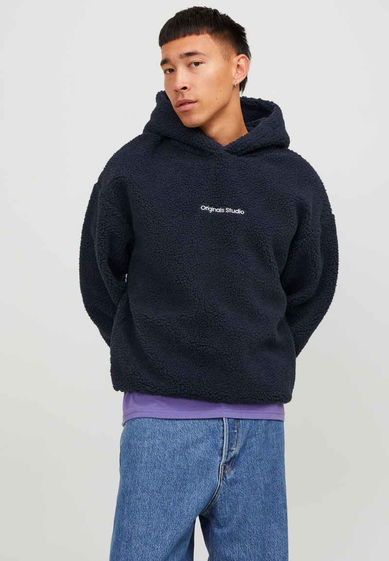 Navy blue fleece hoodie with a textured surface and a kangaroo pocket, embroidered "Originals Studio" logo on the chest. Worn over a purple shirt.