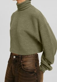 Olivově zelený krátký rolák z texturovaného úpletu, doplněný hnědými džíny s vysokým pasem, které mají měděné kování a vybledlé detaily.