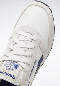 Reebok Classic sneakers med en krämfärgad övre i canvas, mockadetaljer, vita snören och en blå logotyp med geometrisk design. Svart sula.