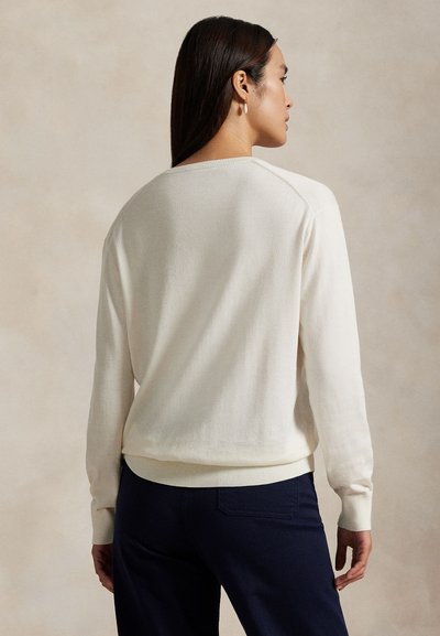 Põhjataustaga naine, kellel on pikad tumedad juuksed, valge pika varrukaga kampsun ja tumedad püksid, näidatud tagant.