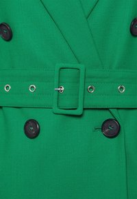 Close-up van een groene stoffen trenchcoat met een doppelsluiting, zwarte knopen, verstelbare riem met metalen hardware en versterkte stiksels.