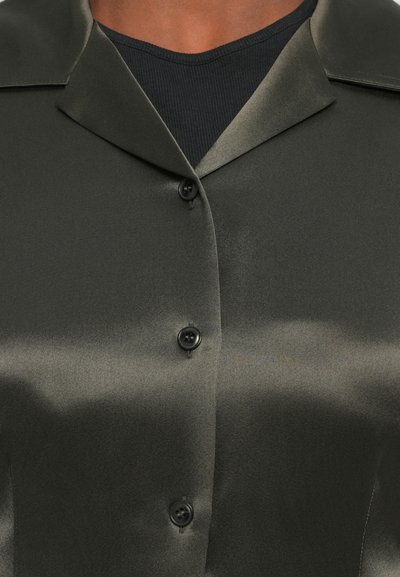 Camicia di raso verde scuro con una texture liscia, dotata di un colletto a intaglio e bottoni neri lungo la parte anteriore, indossata sopra una maglietta a coste scura.