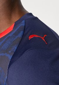 Morska sportska majica s teksturiranim uzorkom, crvenim okruglim izrezom i crvenim Puma logotipom na ramenu. Materijal je gladak i prilagođen tijelu.