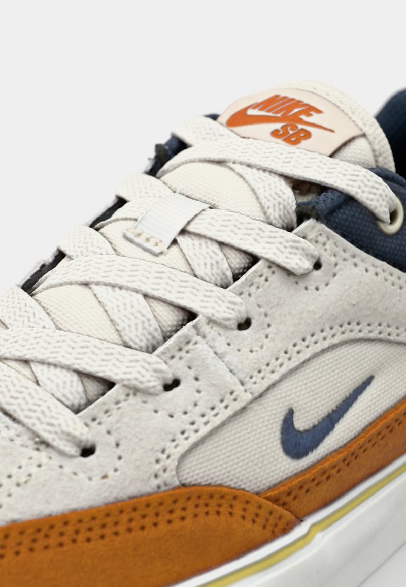 Nike SB Sneaker mit einem cremefarbenen Wildleder-Obermaterial, orangen Wildleder-Akzenten, navy Details und weißen Schnürsenkeln. Auffälliges durchlöchertes Design und Markenpatch.