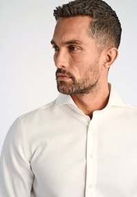 Camicia bianca con colletto strutturato e texture liscia. Presenta bottoni in perla lungo la patta anteriore e una silhouette aderente.