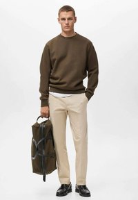 Bruin coltrui, beige broek en zwarte schoenen; houdt een groene tas vast met een zwarte ritssluiting en accenten. Glad textuur en op maat gemaakte pasvorm.