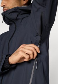 Vandtæt navyjakke med høj krave, lynlåslomme på brystet, teksturerede ærmekanter og sømløs konstruktion for fleksibilitet.