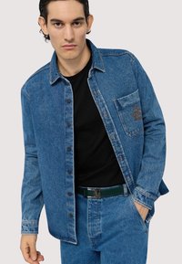 Jasnoniebieska koszula dżinsowa z zapięciem na guziki i kieszonką na piersi; ma kontrastowe przeszycia i teksturowane wykończenie. Nosi się ją na czarnej koszuli.